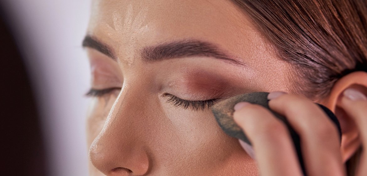 Junge Frau verblendet Concealer mit einem Beauty-Schwämmchen unter ihren Augen.